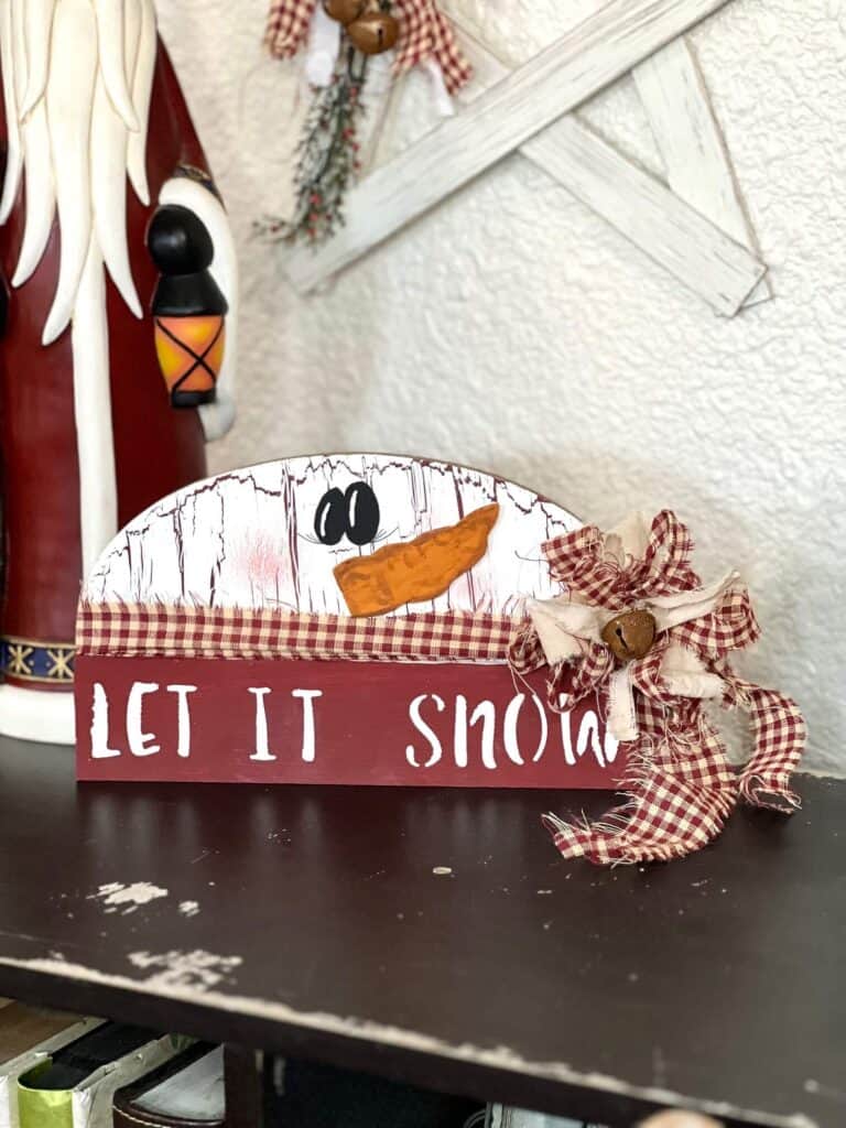 Dollar Tree half wood snowman with white paint and crackle paint underneath, cardboard carrot nose, homespun fabric scarf and bow, and the bottom portion is dark red and say let it snow.