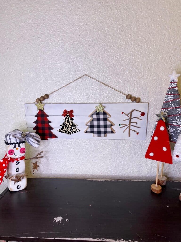 The completed Christmas Tree decor project hanging on the wall with a Snowman to the left and yarn wrapped standing Christmas trees to the right.