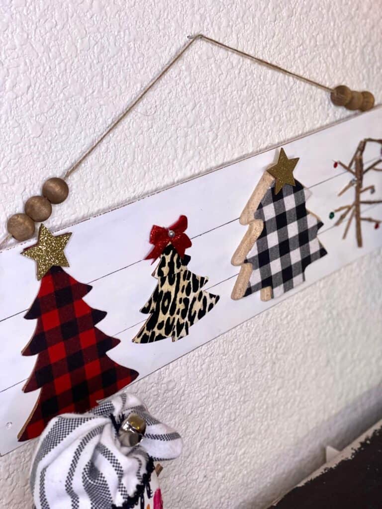 Mixed Christmas tree sign on a long wooden faux shiplap background with red and black buffalo check christmas tree, black and white buffalo check tree, leopard print tree, and a charlie brown christmas tree with a partial wood bead hanger displayed on the wall for DIY Christmas decor.