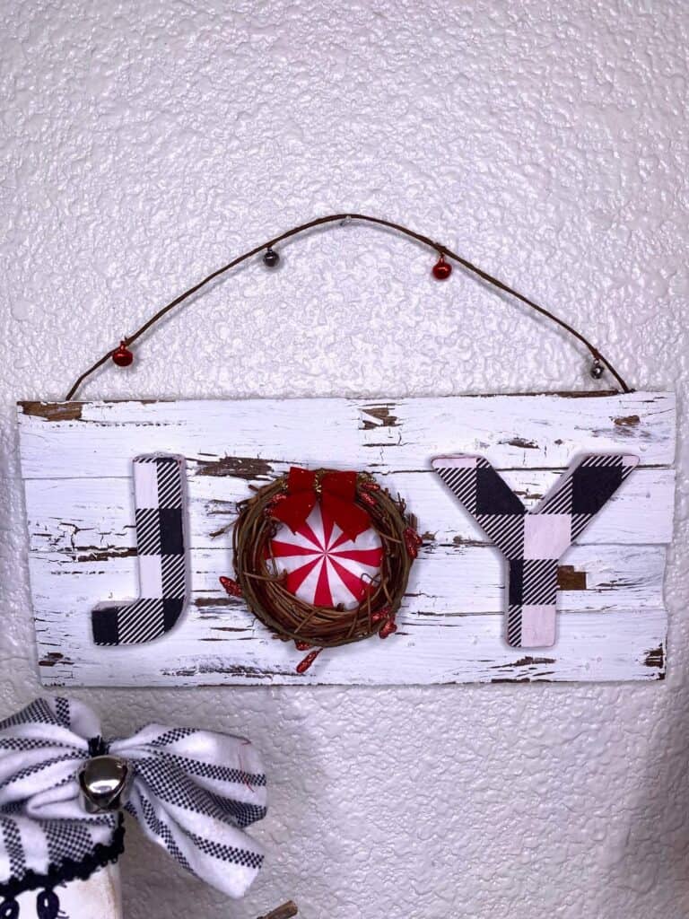 DIY JOY Christmas Sign on a white distressed chippy background with the O as a grapevine wreath with a peppermint napkin inside and a berry wired garland hanger.
