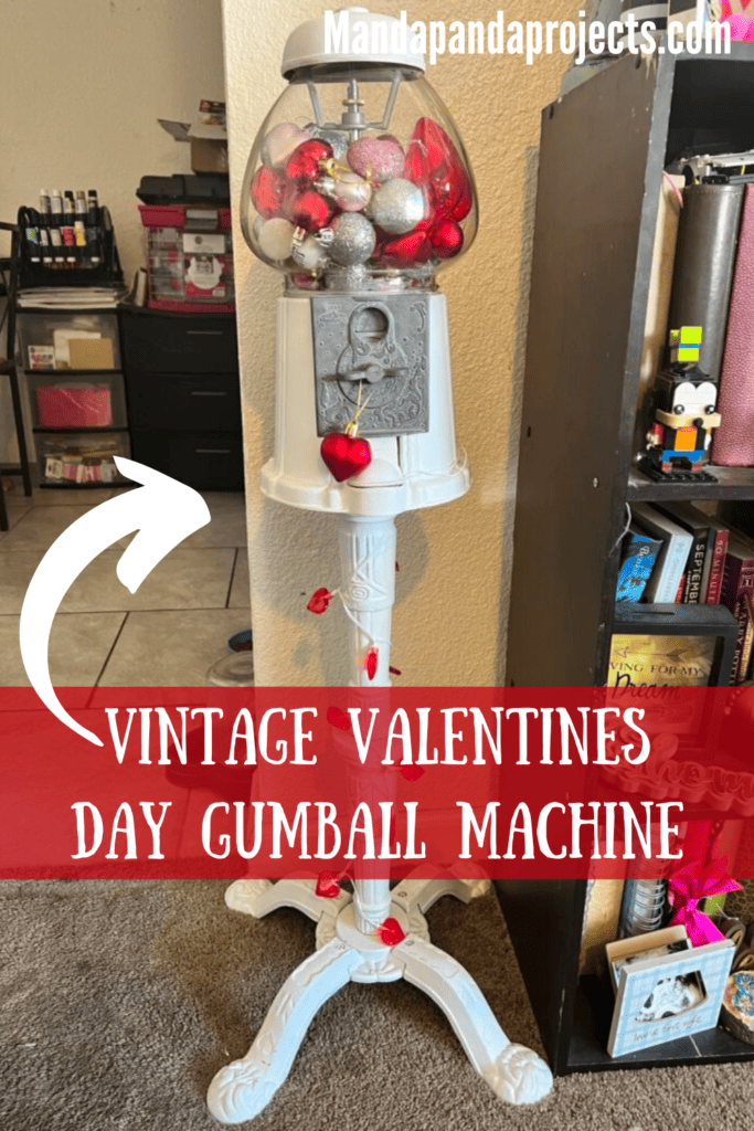 Vintage thrift store gumball machine that has been repainted white and decorated for Valentines Day with red, pink, and silver heart shaped ornaments inside, and a red heart string of lights around the stand. Fun unique Valentines Day heart decor.