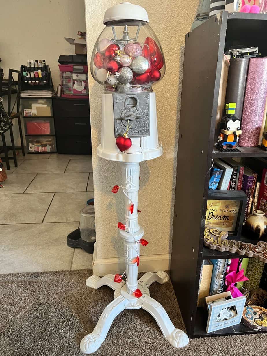 Vintage thrift store gumball machine that has been repainted white and decorated for Valentines Day with red, pink, and silver heart shaped ornaments inside, and a red heart string of lights around the stand. Fun unique Valentines Day heart decor.