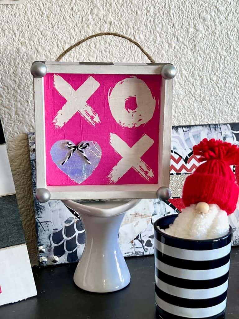 XOXO Valentines Day Napkin Shelf Sitter with a hot pink XOXO napkin that the second O is a silver heart, with a silver half wood bead on each corner and black and whit buffalo check print on each of the 4 sides of the box.