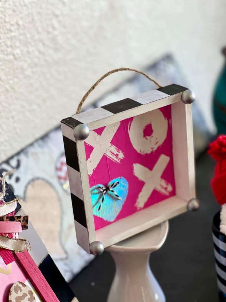 XOXO Valentines Day Napkin Shelf Sitter with a hot pink XOXO napkin that the second O is a silver heart, with a silver half wood bead on each corner and black and whit buffalo check print on each of the 4 sides of the box.