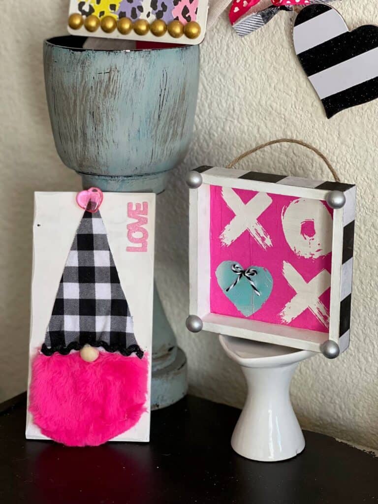 Gnome with a hot pink beard and black and white buffalo check hat, next to the XOXO napkin box frame sitting on a bookcase.