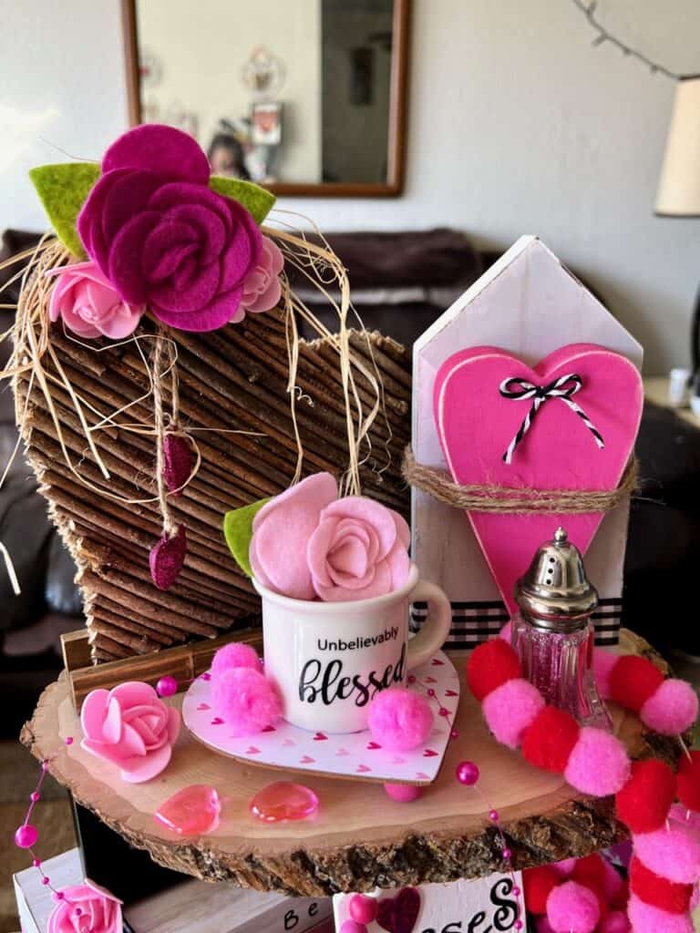 Top tier of the tray with a stick heart, mini mug that says blessed, and wood house with pink chunky heart. Mini roses and pink and red pom poms throughout the tray.