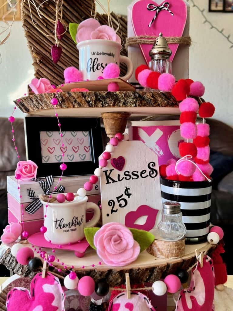 Middle tier of the tray with  Kisses wood tag with lips, a mini mug that says thankful, a pink and white bookstack, and a black and white striped cup. Pink felt rose in the front.