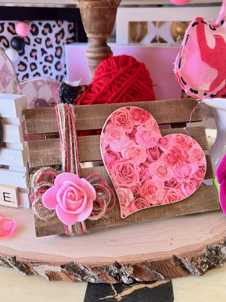Mini wood pallet with a pink floral heart and pink and whit twine with a pink foam rose to the left.