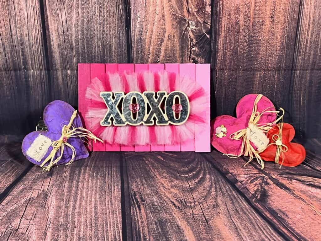 A purple kraft paper heart on the left with a pink and a red kraft paper heart on the right and the completed pink ombre XOXO sign in the middle with a faux wooden background.