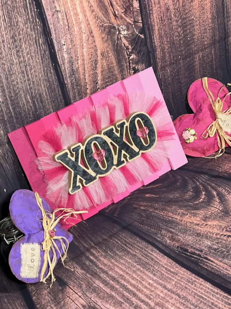 A purple kraft paper heart on the left with a pink kraft paper heart on the right and the completed pink ombre XOXO sign in the middle with a faux wooden background.