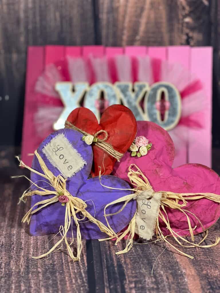 Kraft Paper Valentine Hearts made with brown paper bag, one purple, one red, and one pink, with raffia bow around the center and a white patch and hangtag with the word "love".