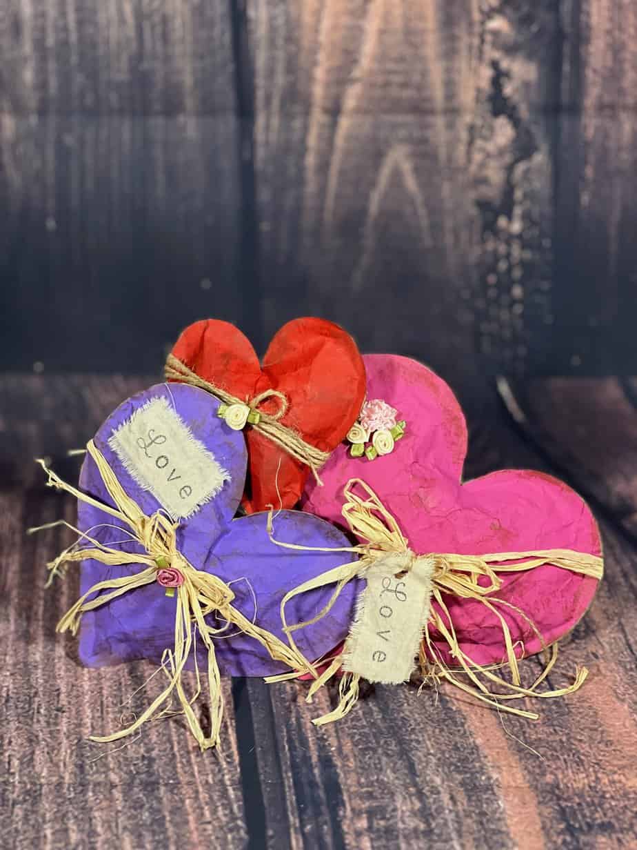 Kraft Paper Valentine Hearts made with brown paper bag, one purple, one red, and one pink, with raffia bow around the center and a white patch and hangtag with the word "love".
