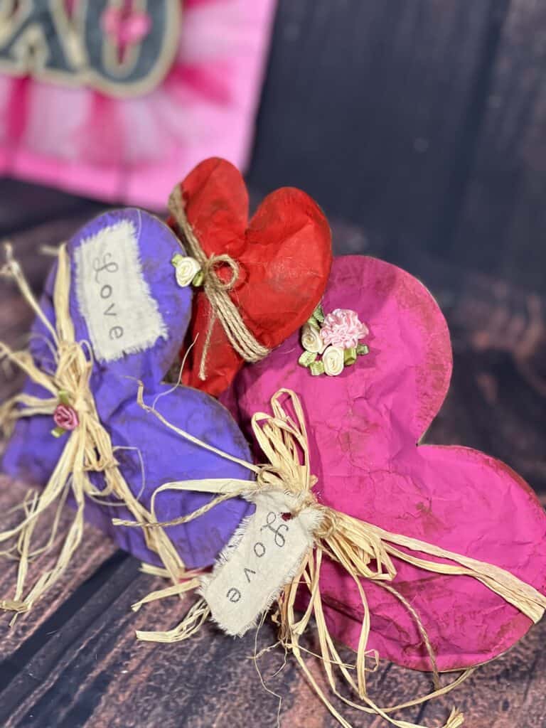 Kraft Paper Valentine Hearts made with brown paper bag, one purple, one red, and one pink, with raffia bow around the center and a white patch and hangtag with the word "love". 