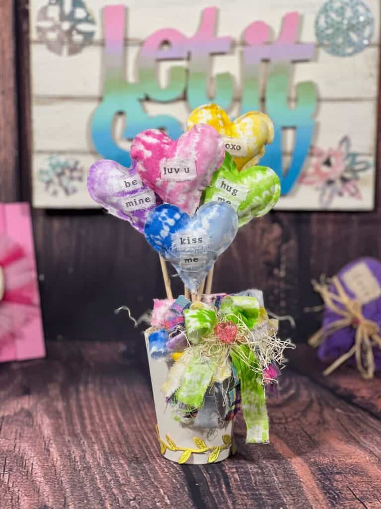 Stuffed Fabric Valentines Day Heart Bouquet with Dollar tree rainbow color tye dye fabric to look like a conversation heart bouquet with valentine phrases stamped on each heart and a big messy colorful bow in a white jar.