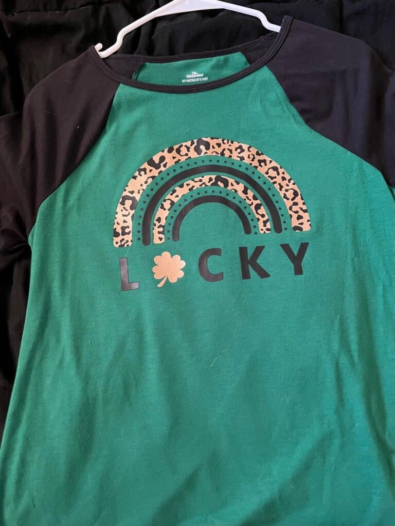 A green shirt with black sleeves with a Leopard print and black rainbow, underneath is the word LUCKY with a shamrock as the "U".