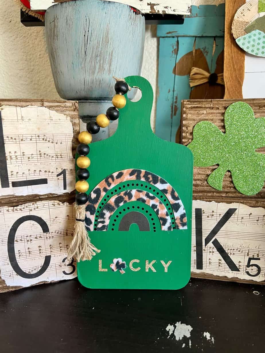 LUCKY Leopard Rainbow Cutting Board for St. Patricks Day with a black and leopard rainbow, the word LUCKY where the "U" is a shamrock, and a gold and black beaded garland on top. Easy to make DIY St Patty's Day decor.