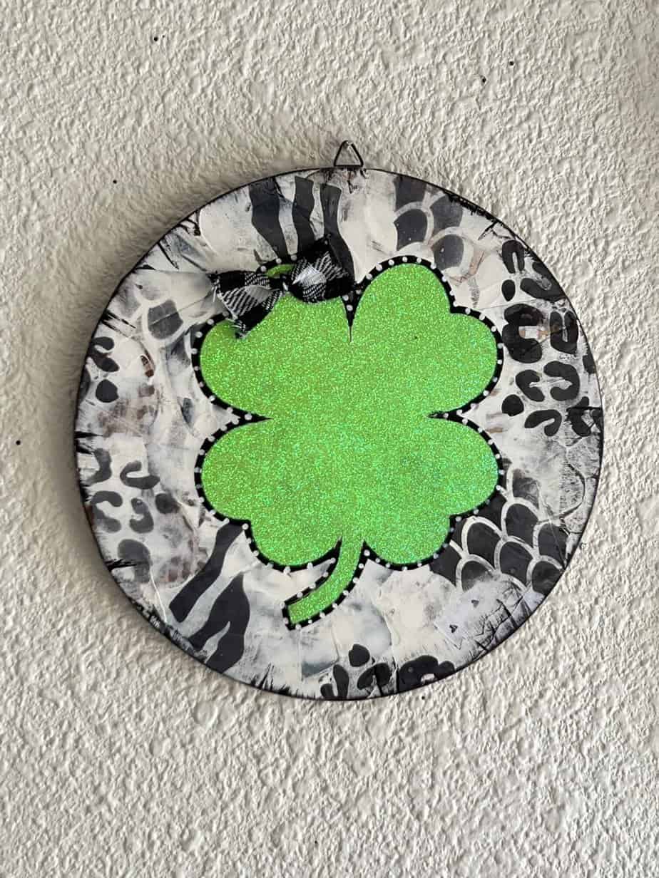 Mixed Media 4 leaf clover with black and white patchwork and animal print stencil background, a glitter light green 4 leaf clover with a small buffalo check bow for St. Patricks Day Decor made on a Dollar Tree wood round.