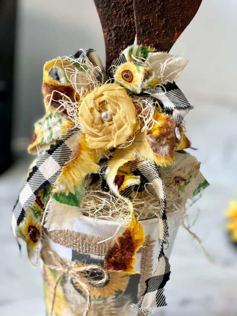 A scrappy fabric bow with sunflower, buffalo check, and white muslin fabric with a yellow fabric rose and a pearl bead center.
