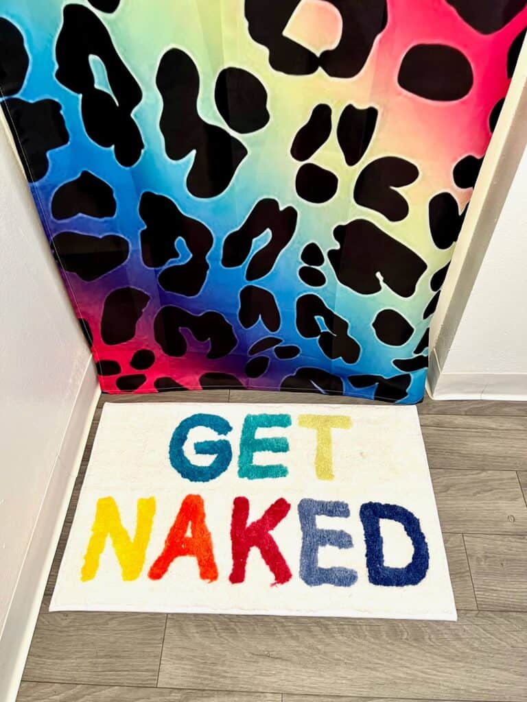 A colorful bath mat on the ground that says "Get Naked" on rainbow colors.
