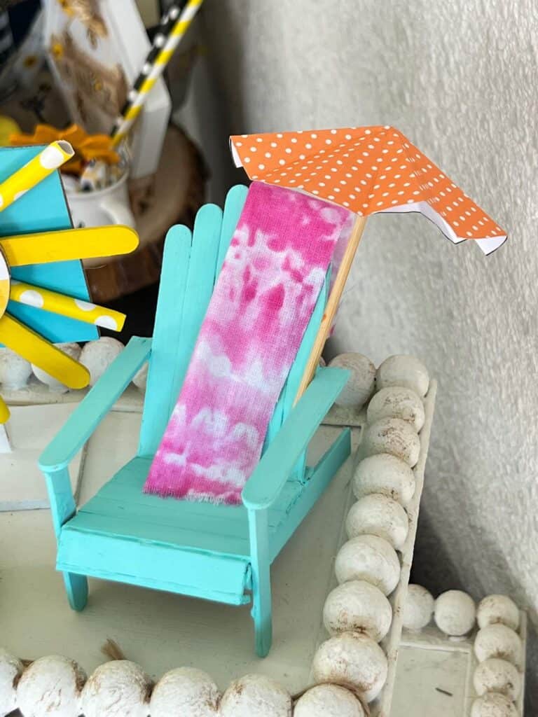 Popsicle stick beach chair with a beach umbrella and a fabric pink towel.