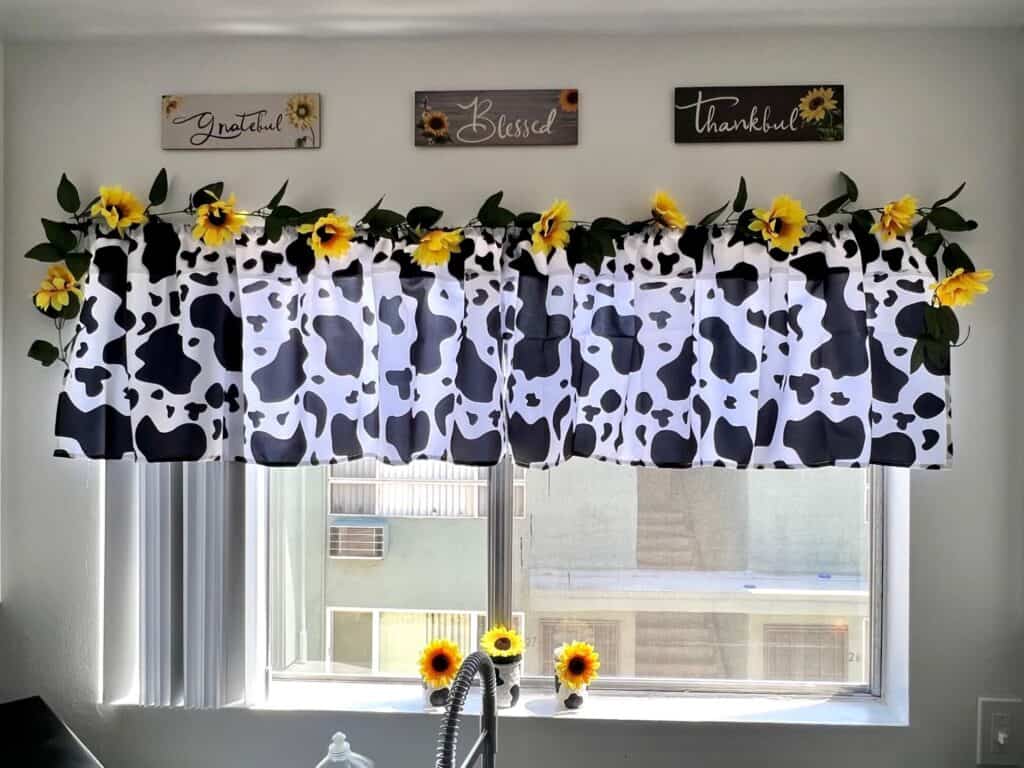 Big kitchen window with a cow print valence and a string of sunflowers on top. Above the window is 3 wall plaques that say Grateful, Thankful, and Blessed. 3 cow print mason jars with sunflowers on top, in the windowsill.