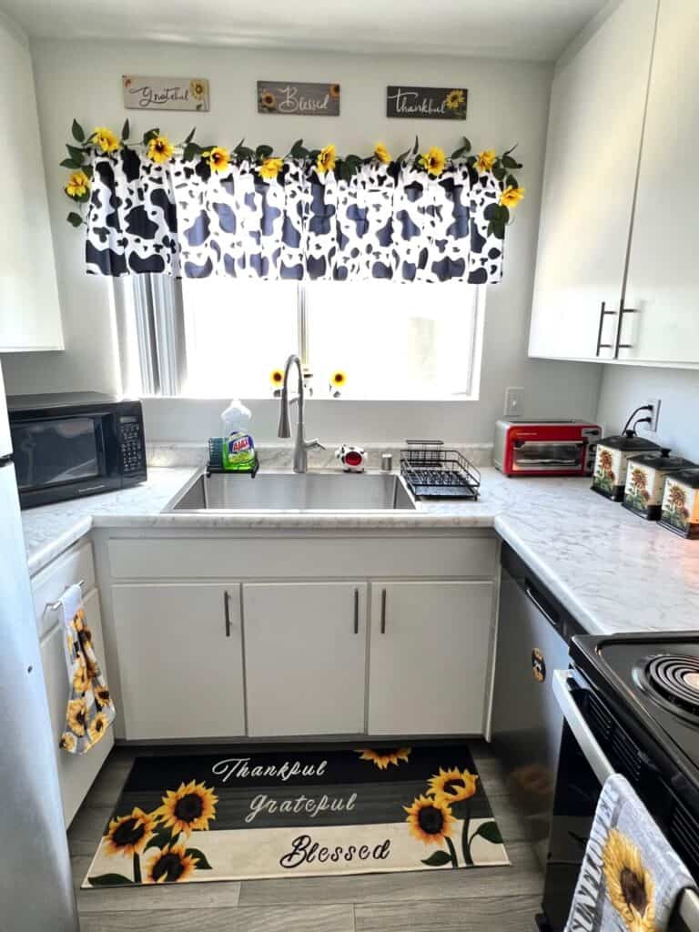 Big kitchen window with a cow print valence and a string of sunflowers on top. Above the window is 3 wall plaques that say Grateful, Thankful, and Blessed. Small cow sponge holder on the sink and a sunflower rug on the floor.