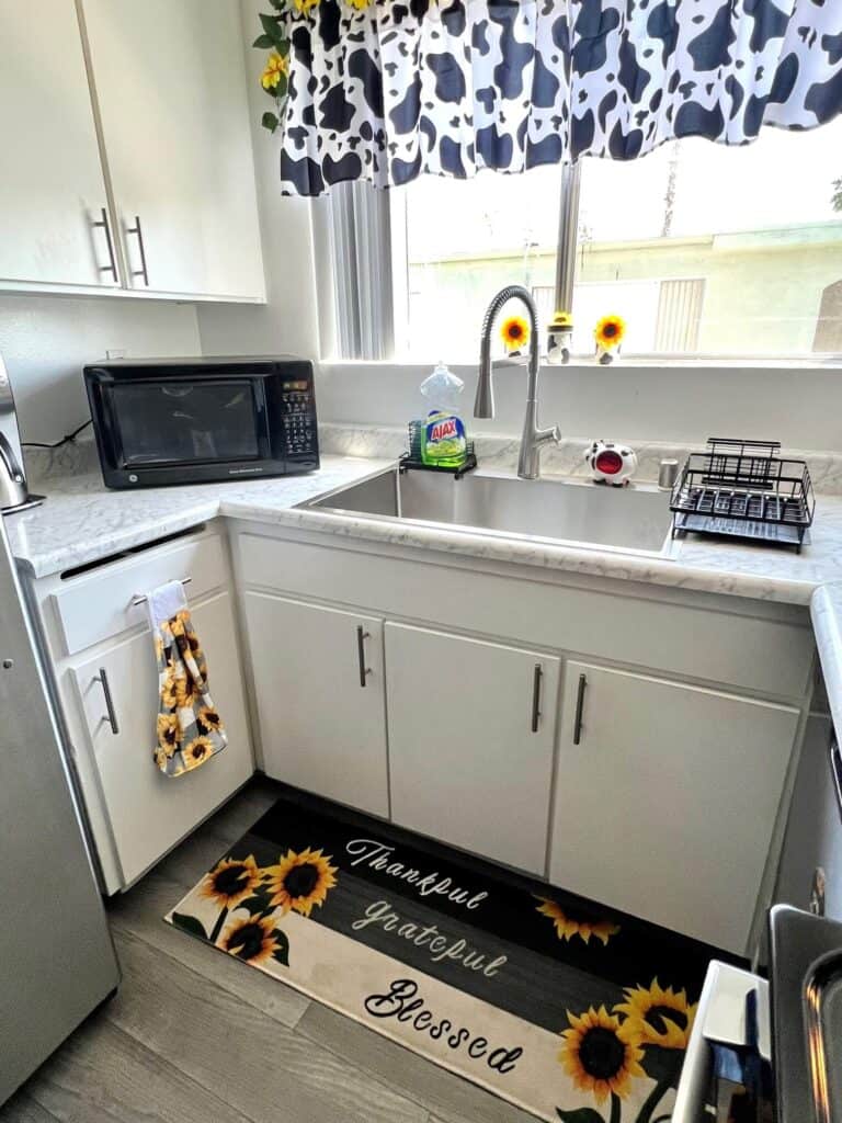 The sink and kitchen counter with a small cow sponge holder on the sink, a Sunflower kitchen rug on the floor and a buffalo check sunflower hand towel hanging from the drawer handle.