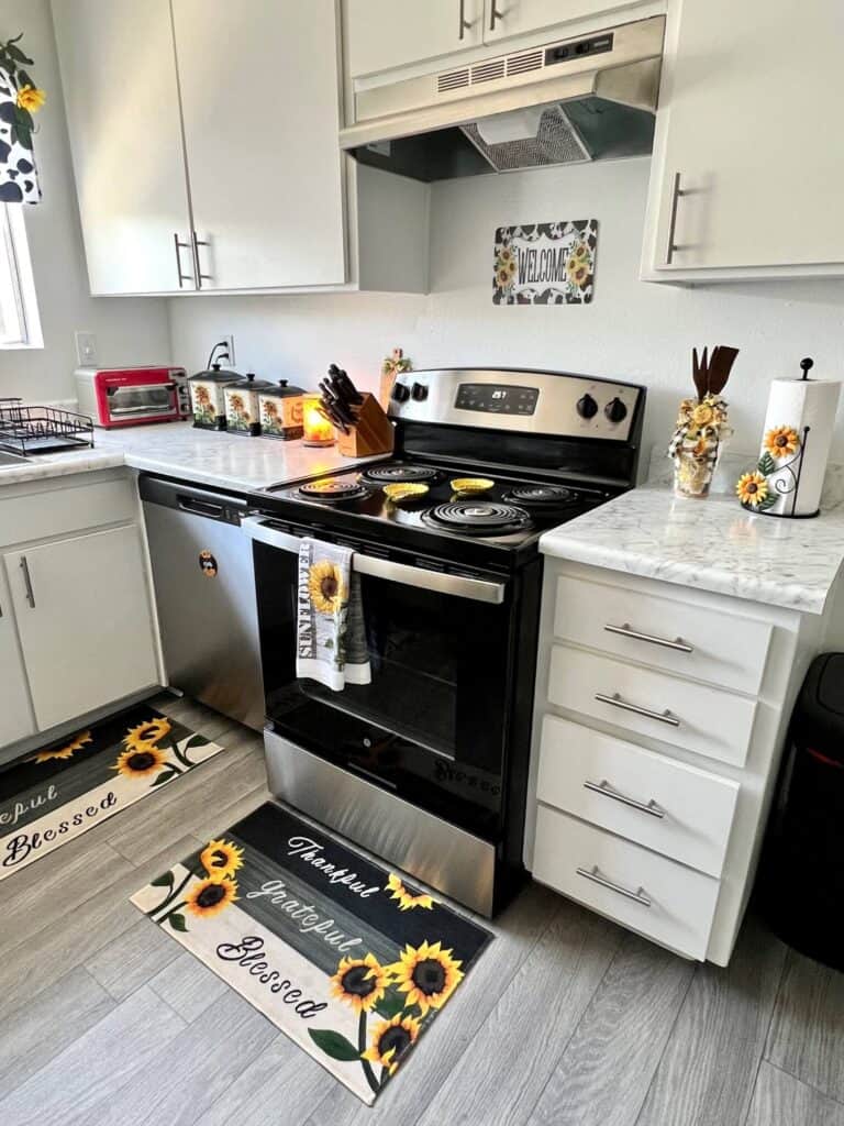Stove with sunflower decor, spoon rests, and hand towel. Kitchen Counters with Sunflower paper towel holder, canisters, and rusted utensil jar. Floor with Sunflower kitchen mats.