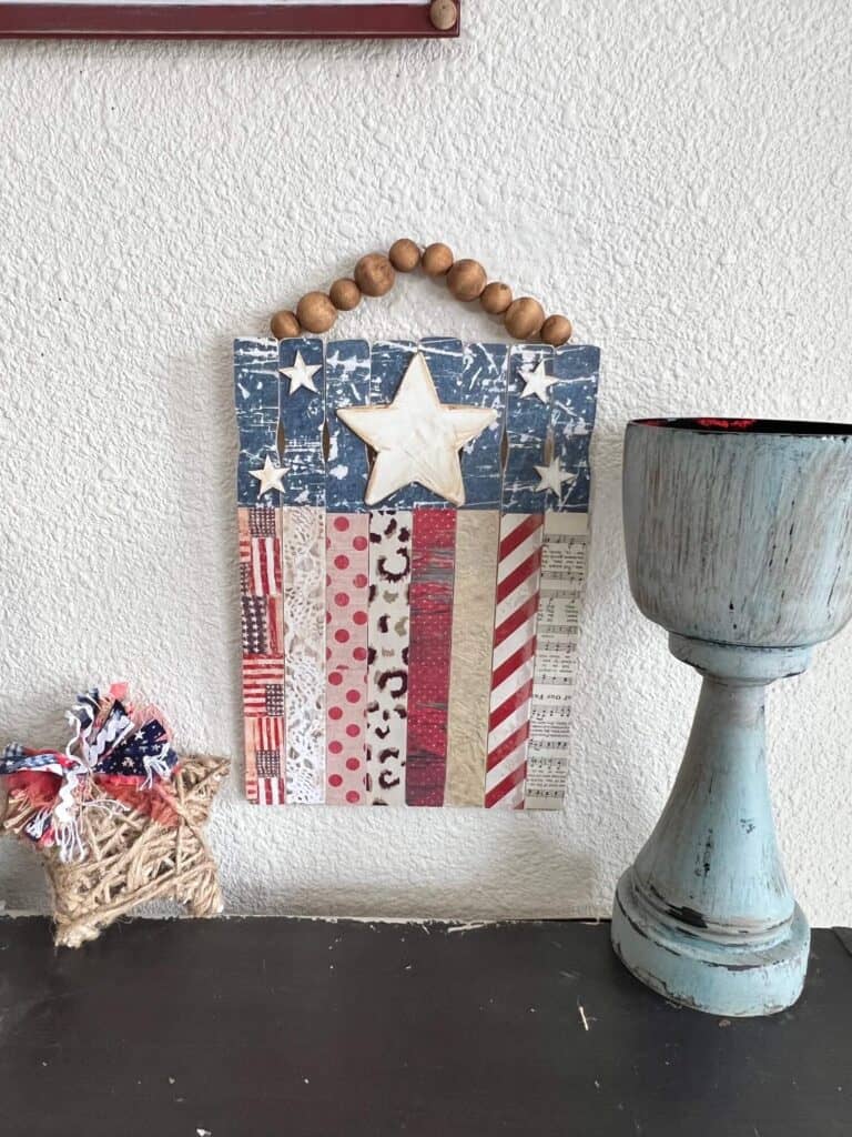 The completed paint stick american flag hanging on the wall next to a chunky candlestick and a twine patriotic star.