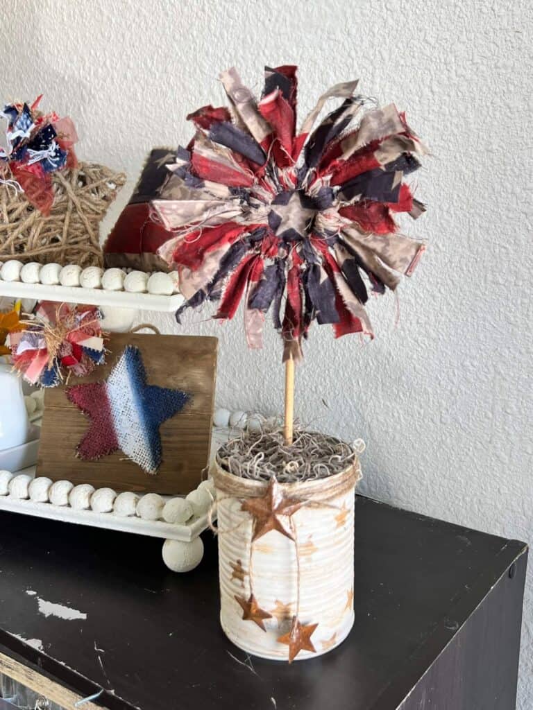 Patriotic Fabric Ring Flower with a recycled tin can base painted white with antique wax stars, twine and a rusty star made with red white and blue American flag fabric and dollar tree wood ring center. Rustic 4th of july DIY Decor.