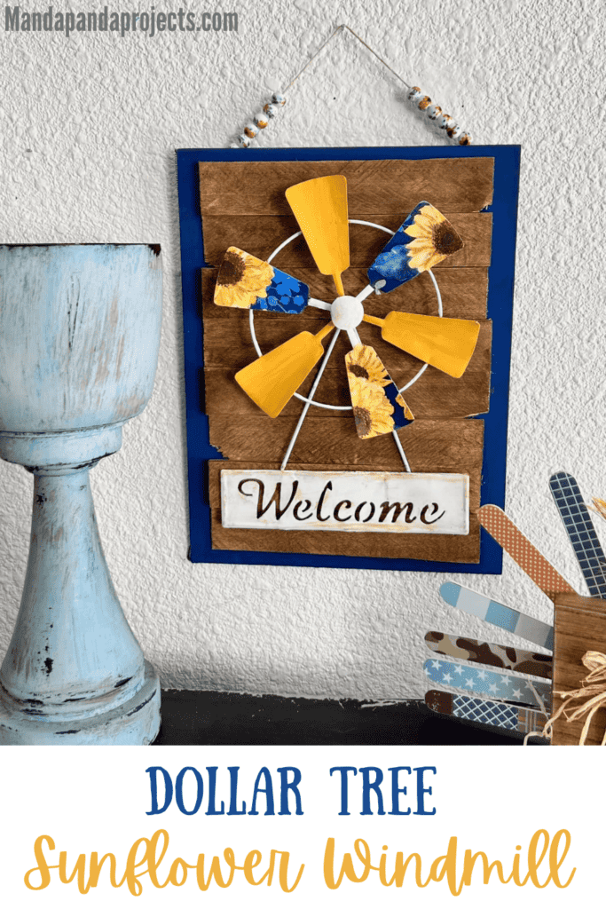 Dollar Tree Sunflower Windmill welcome sign with a blue and sunflower napkin decoupaged onto the windmill, a blue background with brown stained shims layered on top, and a ceramic sunflower bead hanger for DIY budget decor hanging on the wall.
