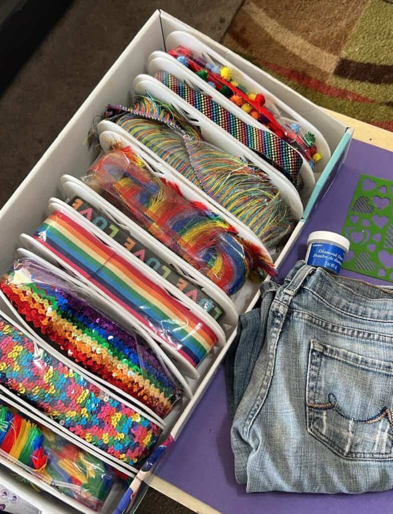 Tray of rainbow trims and pom poms and a pair of jeans.
