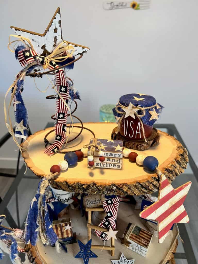 Top tier of the tray with a rusty patriotic star, mini bookstack, wood bead garland, and USA patriotic jar.