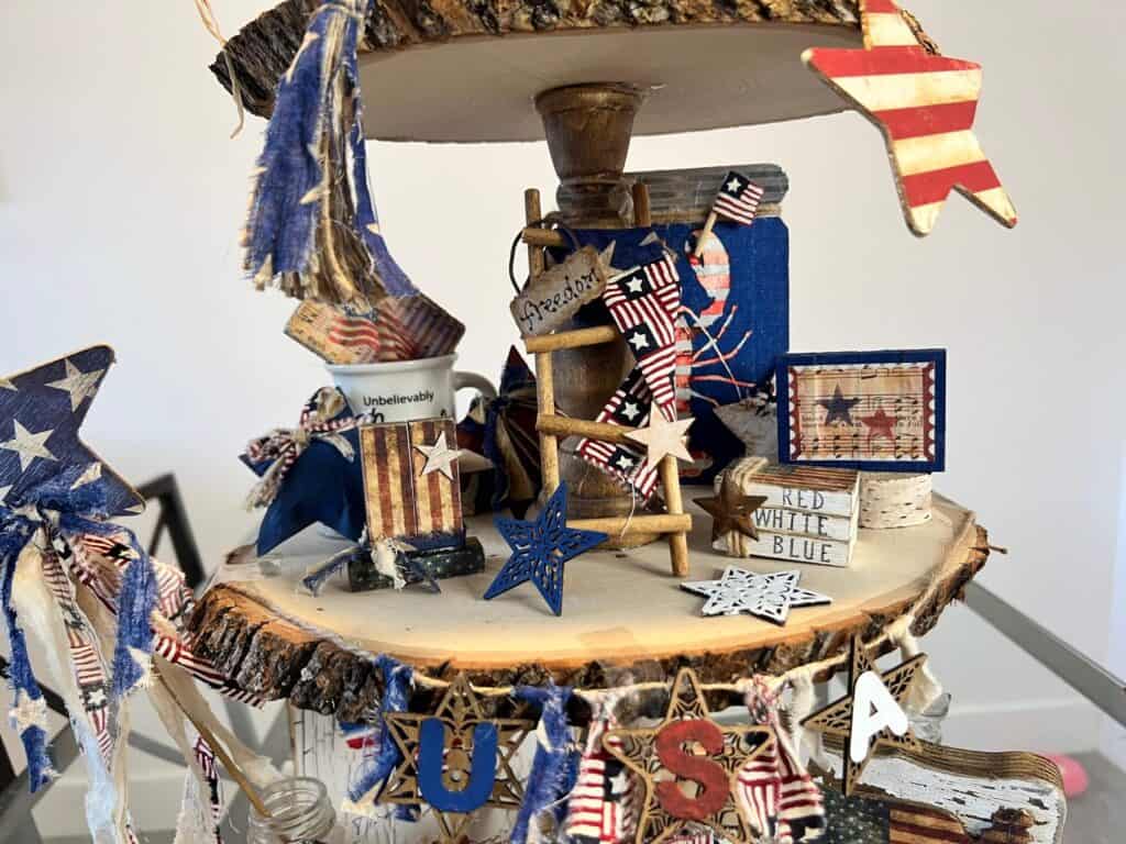 Middle tier of the tray with a Patriotic Ladder, Uncle Sam hat, mini book stack, and Patriotic Lobster Mason Jar. USA star banner across the bottom of the wood round tier.
