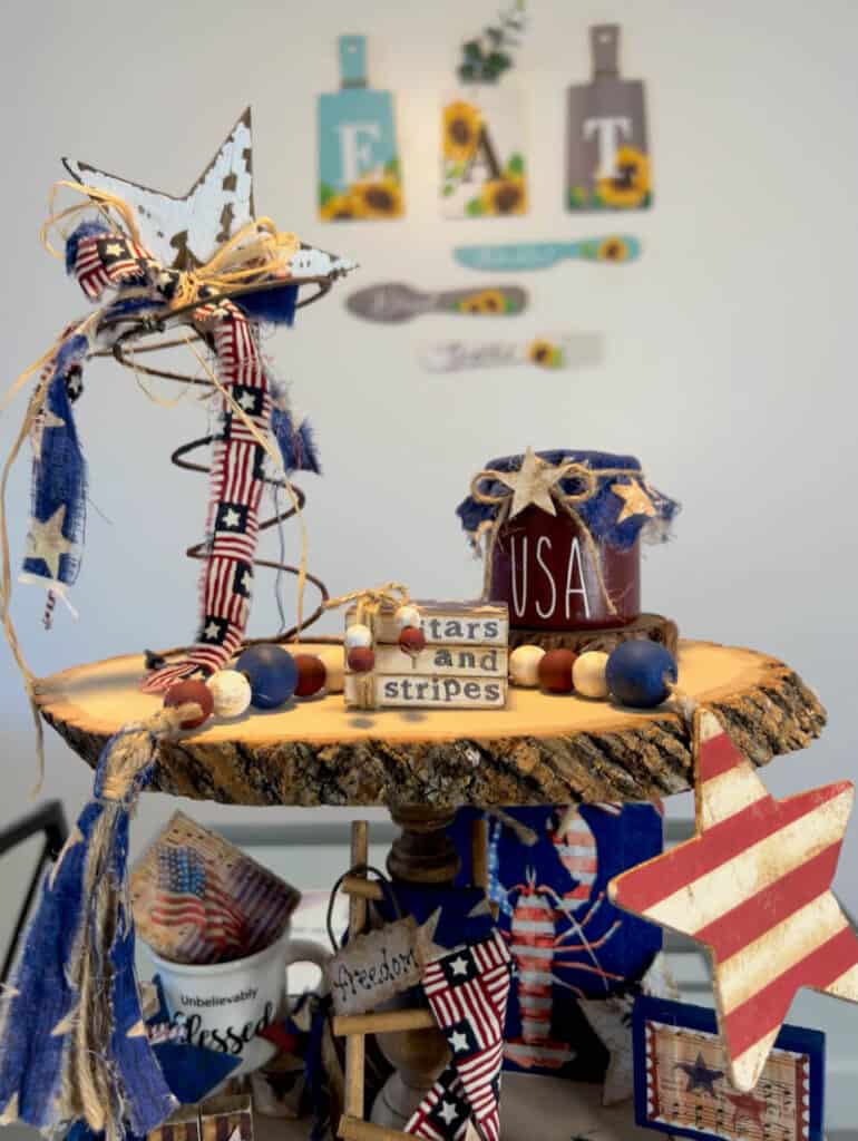 Top tier of the tray with a rusty patriotic star, mini bookstack, wood bead garland, and USA patriotic jar.