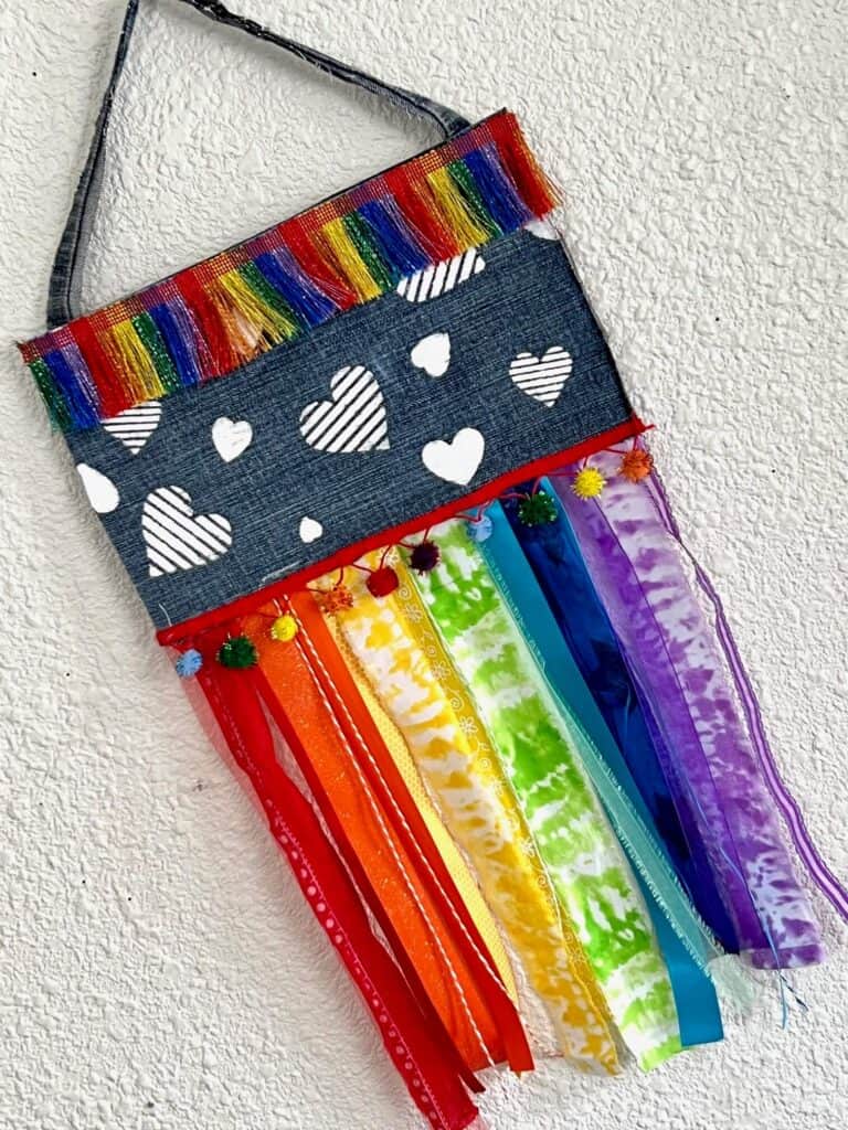 DIY Denim Rainbow Rag Flag with rainbow fabric hanging strips, multicolor trim and pom poms, and white stenciled hearts to show your Pride for the month of June and celebrate that Love is Love.