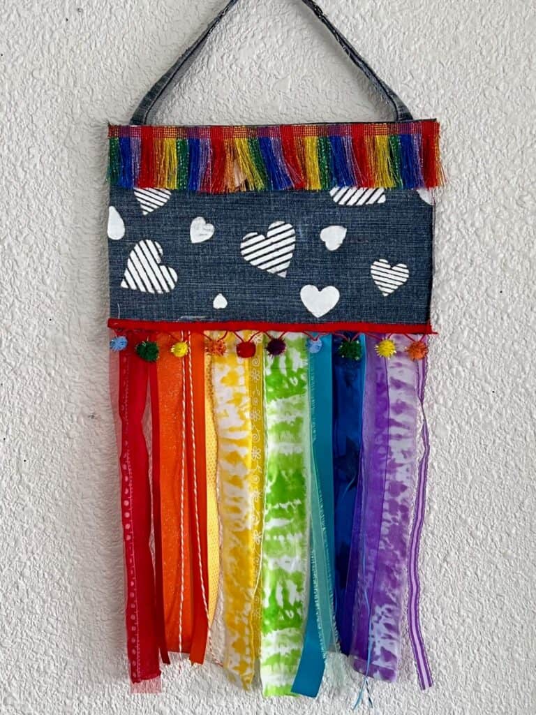 DIY Denim Rainbow Rag Flag with rainbow fabric hanging strips, multicolor trim and pom poms, and white stenciled hearts to show your Pride for the month of June and celebrate that Love is Love.