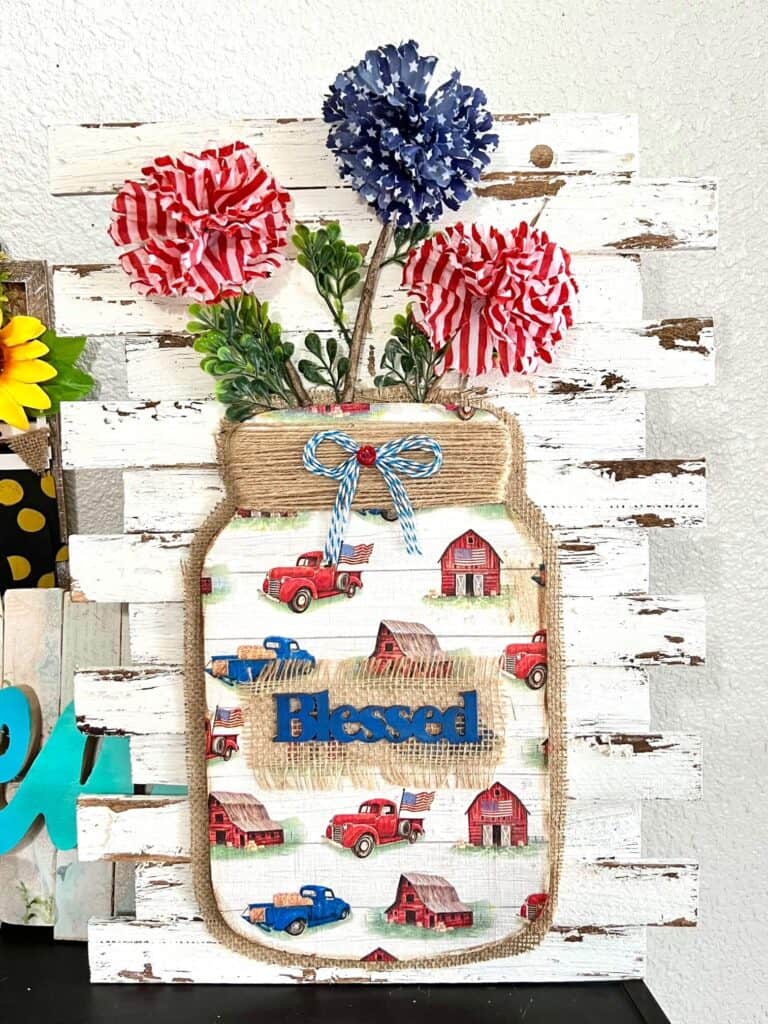 Patriotic Floral Mason Jar with wood cutout, vintage red, white, and blue truck and barn, stars and stripes dollar tree flowers on a white chippy shim background to decorate for the 4th of July or memorial day.