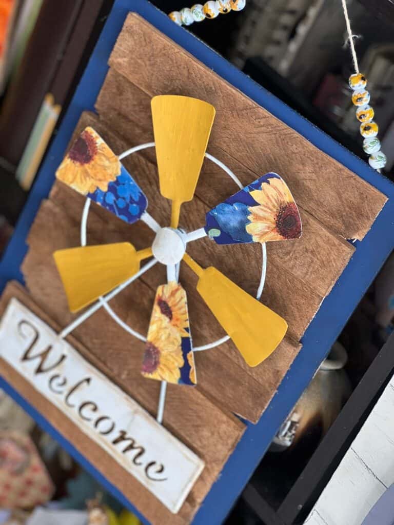 Dollar Tree Sunflower Windmill welcome sign with a blue and sunflower napkin decoupaged onto the windmill, a blue background with brown stained shims layered on top, and a ceramic sunflower bead hanger for DIY budget decor hanging on the wall.
