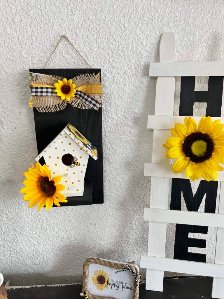 The sunflower birdhouse hanging on the wall to the left of a sunflower HOME paint stick ladder.