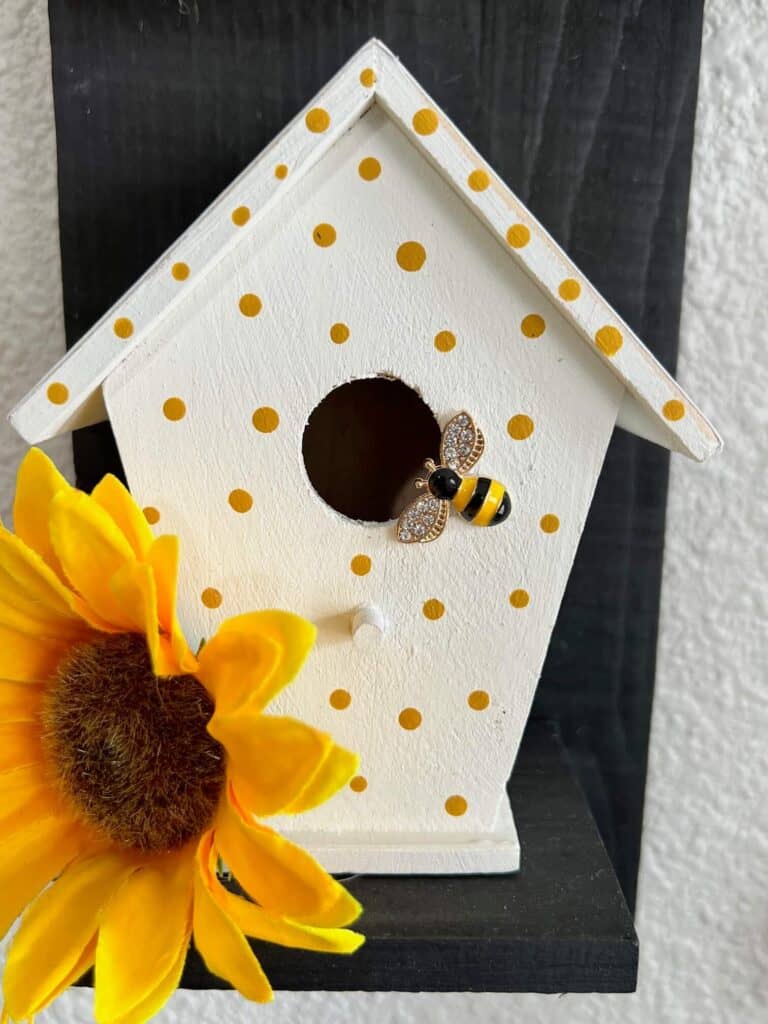 The front of the birdhouse painted white with little yellow polka dots, a rhinestone bumblebee, and a sunflower head.