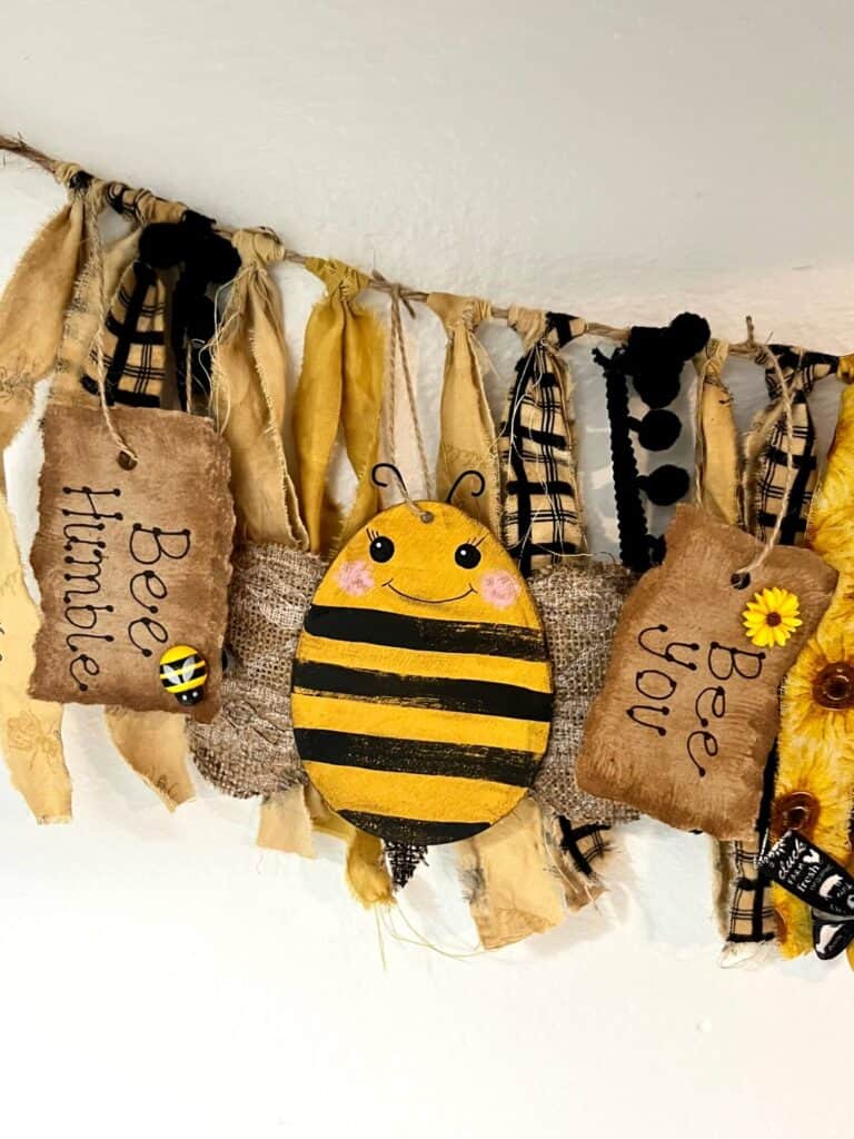 An easter egg bee hanging on the swag banner in between 2 Kraft paper tags, one that says "Bee Humble" and the other says " Bee You", with strips of fabric in between.