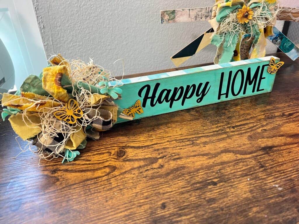 Teal checks, mini butterflies, and a messy bow on a long board that says Happy Home Shelf Sitter made with dollar tree supplies and the words cut out on a cricut for spring summer or everyday DIY home decor.