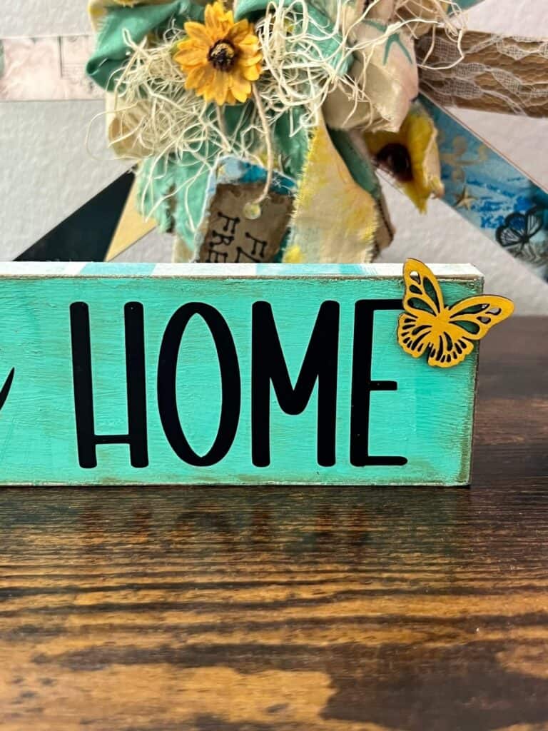 The word Home cut from cricut black vinyl with a small yellow butterfly on the top right and a teal background.