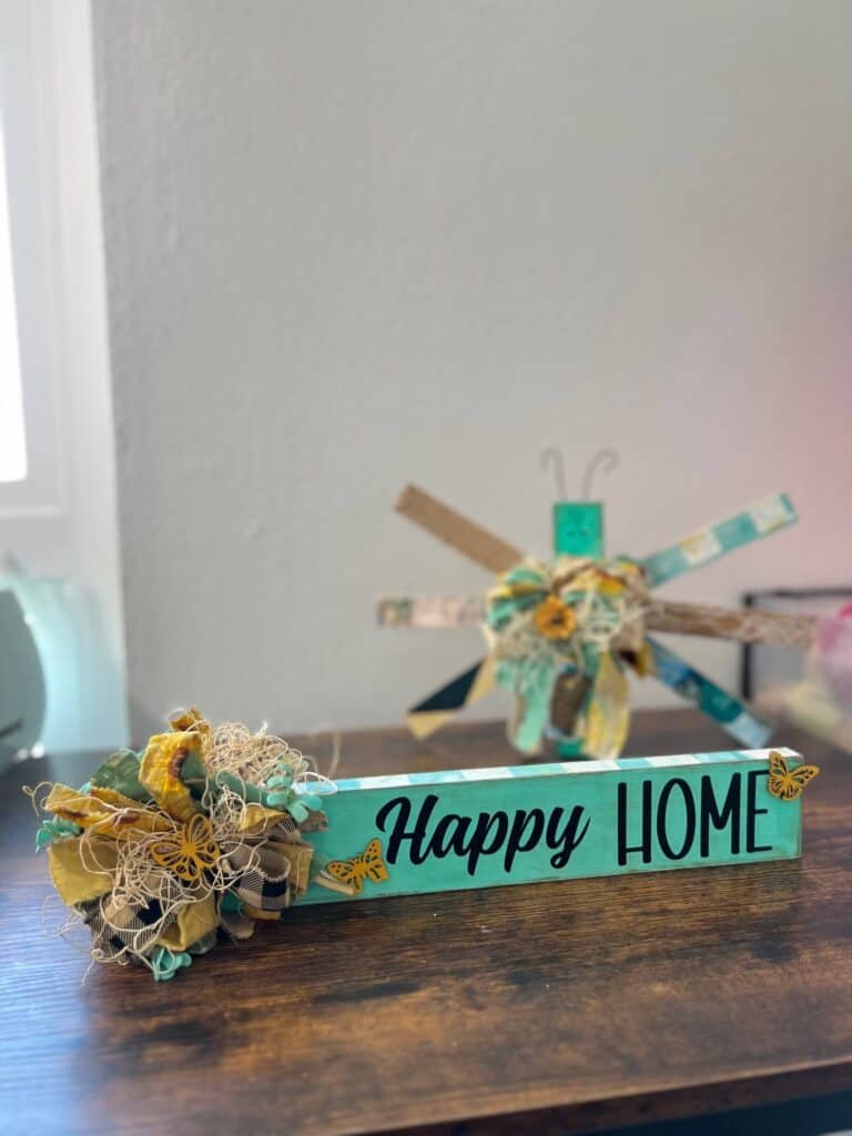 Teal checks, mini butterflies, and a messy bow on a long board that says Happy Home Shelf Sitter made with dollar tree supplies and the words cut out on a cricut for spring summer or everyday DIY home decor.