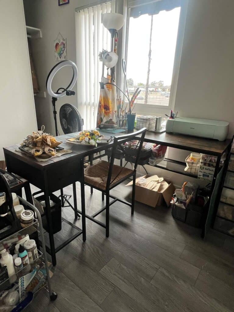 A large L-Shaped desk in front of a big window in a craft room covered in crafts and supplies.