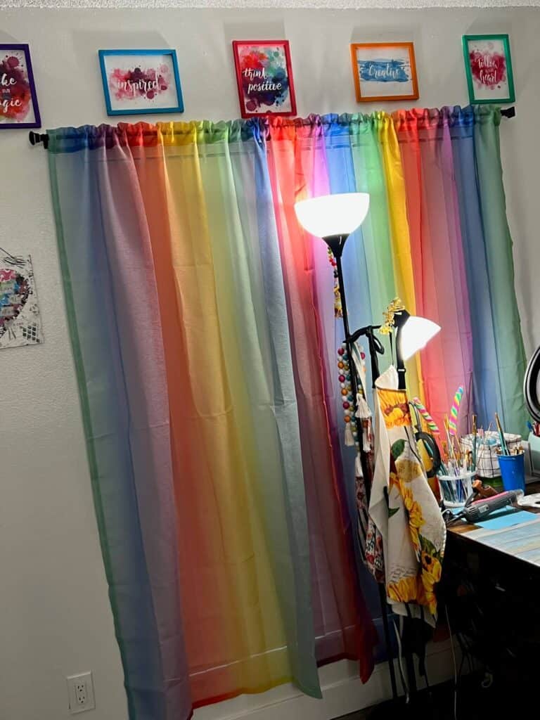 Rainbow curtains hanging in a craft room with a bright light in front and colorful quote designs in frames above the window.