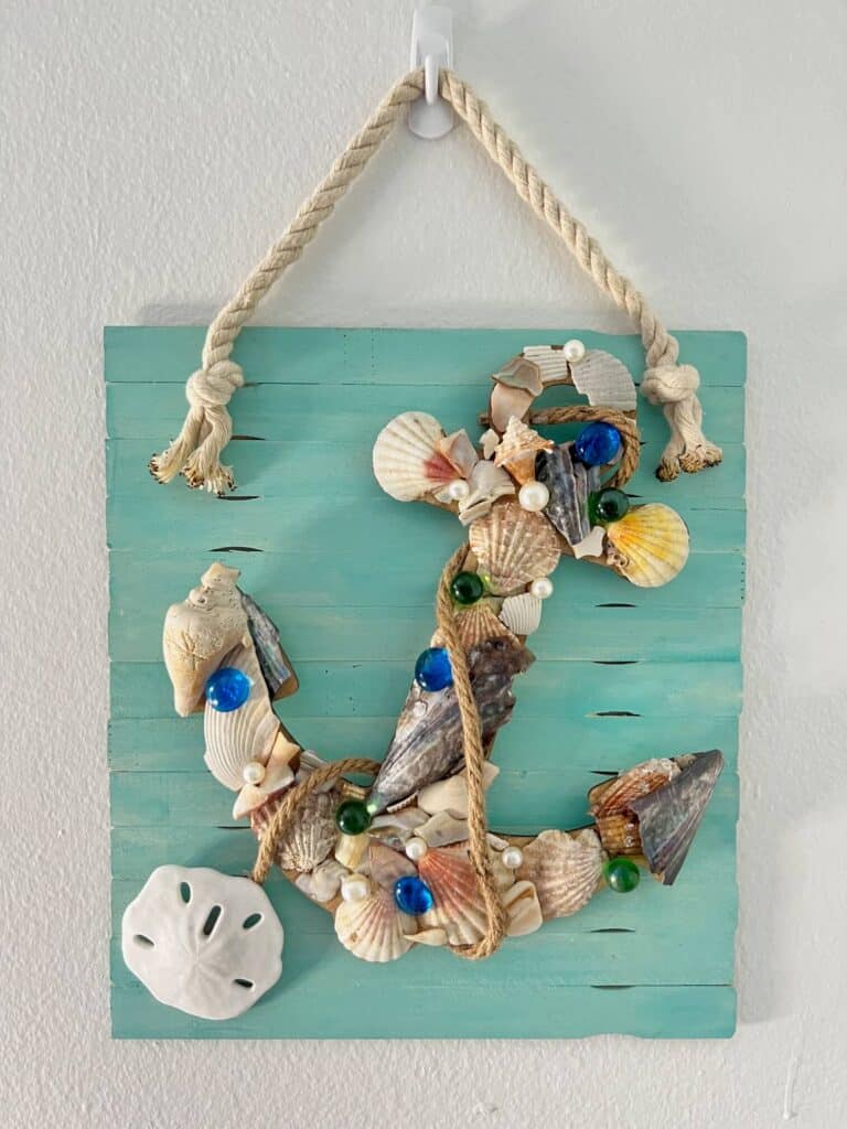 Dollar Tree Seashell Anchor DIY coastal decor with a wood cutout covered in seashells, on a white washed teal background hanging by white nautical rope, with a sand dollar at the bottom.