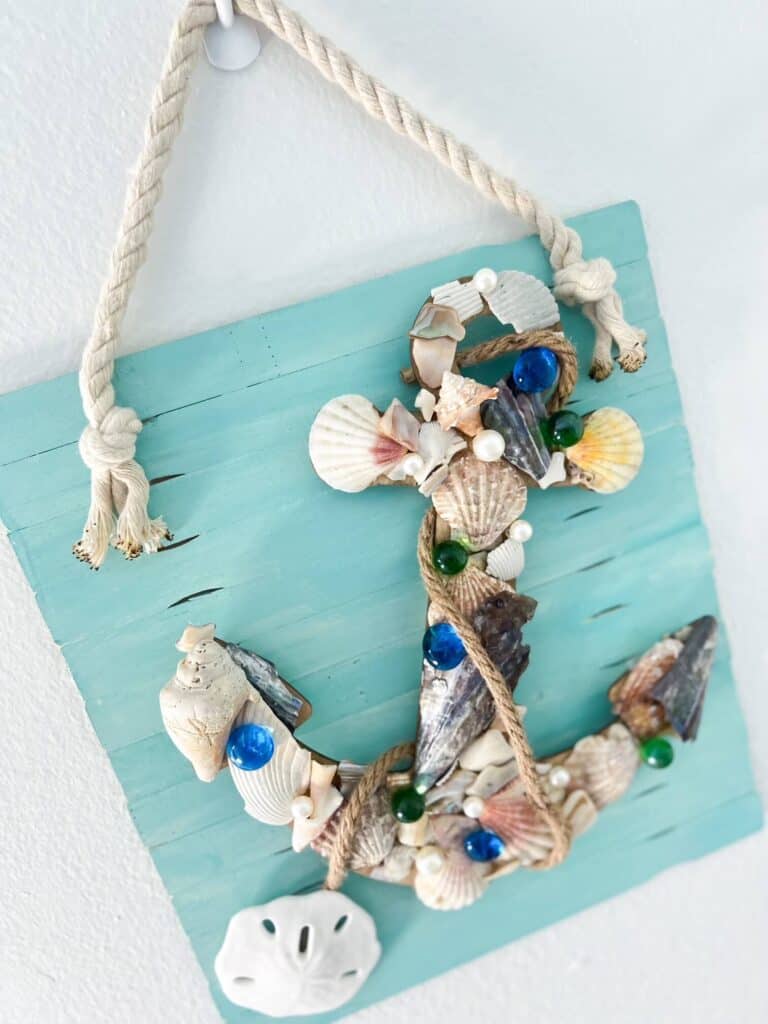 Dollar Tree Seashell Anchor DIY coastal decor with a wood cutout covered in seashells, on a white washed teal background hanging by white nautical rope, with a sand dollar at the bottom.