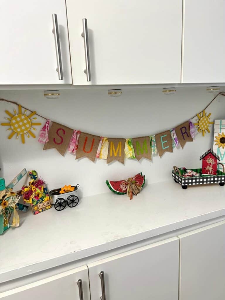 A crafty Summer burlap banner with colorful letters  stenciled on the burlap and fabric strips in between, a yellow sun on each end, hanging on a shelf decorated for summer.